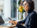 businesswoman with glasses sitting looking at cell phone working on laptop