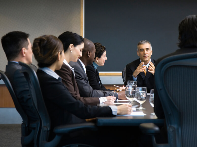 seated businessman leading meeting with seated business women and men listening
